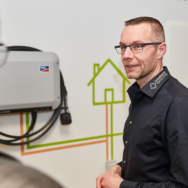 Ein Fachmann steht neben einem an der Wand montierten Wechselrichter mit grünen und orangefarbenen Schaltplänen, die ein Haus darstellen.