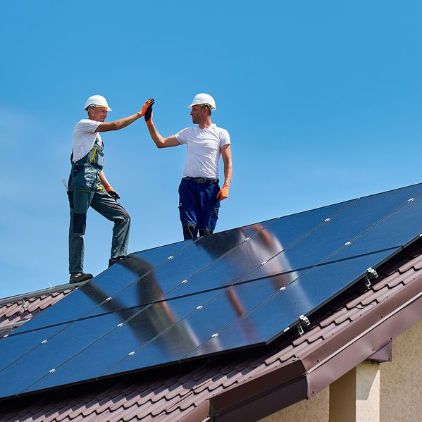 Zwei Bauarbeiter geben sich unter strahlend blauem Himmel auf einem mit Sonnenkollektoren bedeckten Dach ein High Five.