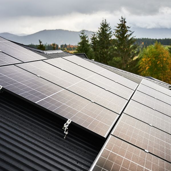 Nahaufnahme von Sonnenkollektoren, die auf einem schwarzen Dach installiert sind, umgeben von grünen Bäumen und fernen Bergen unter einem bewölkten Himmel.